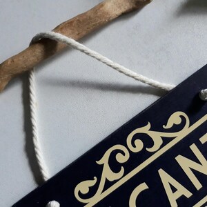 Puede incluir: Una etiqueta rectangular azul marino con detalles dorados y la palabra "CANT" parcialmente visible. La etiqueta est&aacute; suspendida por un cord&oacute;n blanco de una rama de madera marr&oacute;n.