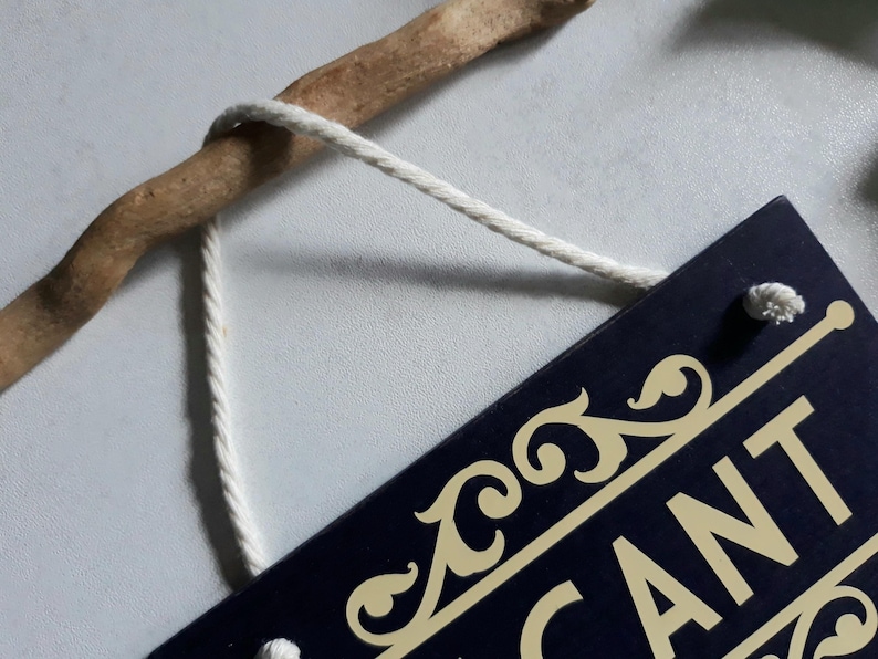 Puede incluir: Una etiqueta rectangular azul marino con detalles dorados y la palabra "CANT" parcialmente visible. La etiqueta est&aacute; suspendida por un cord&oacute;n blanco de una rama de madera marr&oacute;n.