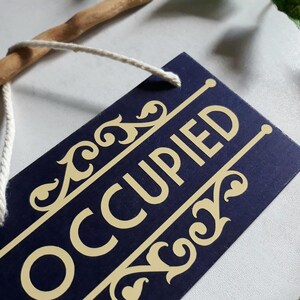 Puede incluir: Un letrero rectangular azul marino con la palabra "OCCUPIED" en letras doradas. El letrero tiene detalles dorados decorativos y est&aacute; sujeto a un palo de madera con una cuerda blanca.