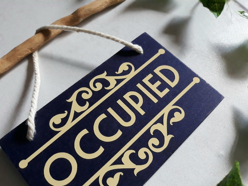 Puede incluir: Un letrero rectangular azul marino con la palabra "OCCUPIED" en letras doradas. El letrero tiene detalles dorados decorativos y est&aacute; sujeto a un palo de madera con una cuerda blanca.