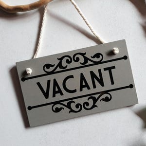 Puede incluir: Un letrero rectangular gris con la palabra "VACANT" en negro, adornado con adornos decorativos. Cuelga de una cuerda blanca. El fondo es blanco con hojas verdes y una rama de madera.