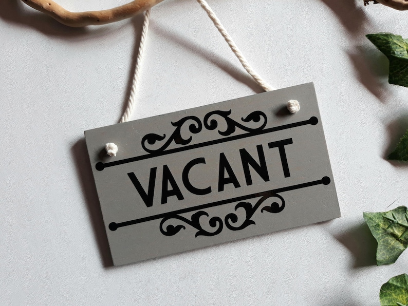 Puede incluir: Un letrero rectangular gris con la palabra "VACANT" en negro, adornado con adornos decorativos. Cuelga de una cuerda blanca. El fondo es blanco con hojas verdes y una rama de madera.