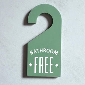 May include: A sage green door hanger with the words "BATHROOM FREE" in white, block letters. The sign has a curved top for hanging and a rectangular bottom. The background is a light gray textured wall.