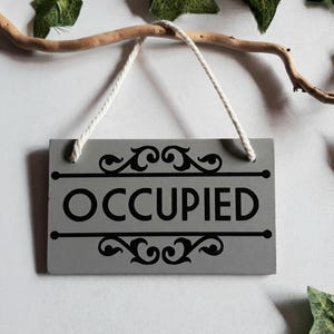 Puede incluir: Un letrero rectangular gris con la palabra "OCCUPIED" en negro, adornado con adornos decorativos. Cuelga de una cuerda blanca, suspendida de una rama. Se ven hojas verdes en el fondo. El letrero indica que una habitaci&oacute;n est&aacute; ocupada.