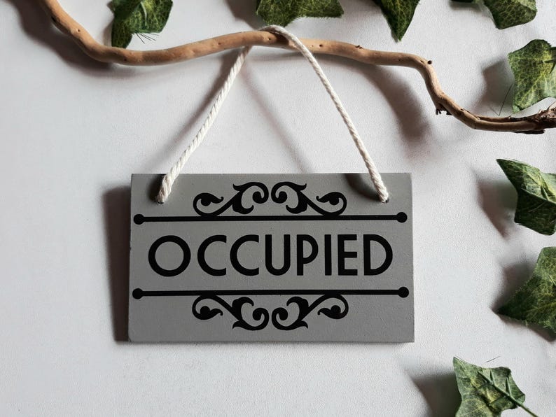 Puede incluir: Un letrero rectangular gris con la palabra "OCCUPIED" en negro, adornado con adornos decorativos. Cuelga de una cuerda blanca, suspendida de una rama. Se ven hojas verdes en el fondo. El letrero indica que una habitaci&oacute;n est&aacute; ocupada.