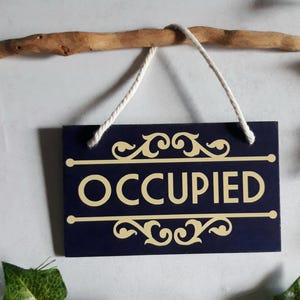 Puede incluir: Un letrero rectangular azul marino con la palabra "OCCUPIED" en letras doradas. El letrero est&aacute; suspendido por un cord&oacute;n blanco de una rama de madera marr&oacute;n. Adornos dorados decorativos est&aacute;n arriba y abajo de la palabra.