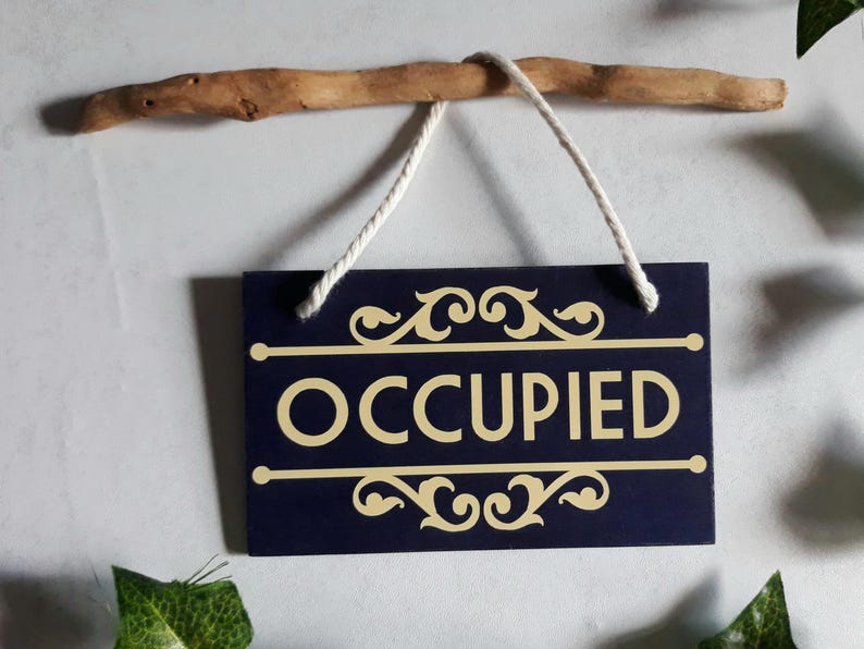 Puede incluir: Un letrero rectangular azul marino con la palabra "OCCUPIED" en letras doradas. El letrero est&aacute; suspendido por un cord&oacute;n blanco de una rama de madera marr&oacute;n. Adornos dorados decorativos est&aacute;n arriba y abajo de la palabra.
