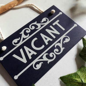 Puede incluir: Un letrero rectangular azul marino con la palabra "VACANT" en blanco, con adornos decorativos. El letrero est&aacute; suspendido por una cuerda blanca y un palo de madera. Hojas verdes en el fondo.