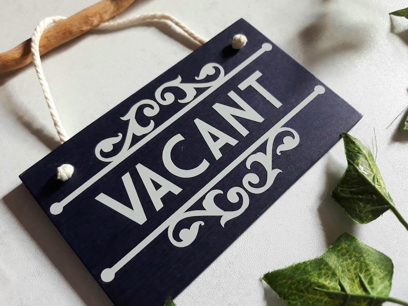 Puede incluir: Un letrero rectangular azul marino con la palabra "VACANT" en blanco, con adornos decorativos. El letrero est&aacute; suspendido por una cuerda blanca y un palo de madera. Hojas verdes en el fondo.