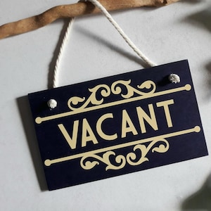 Puede incluir: Un letrero de madera azul marino con la palabra "VACANT" en letras doradas. El letrero tiene adornos dorados decorativos y est&aacute; suspendido por un cord&oacute;n blanco de una rama de madera.