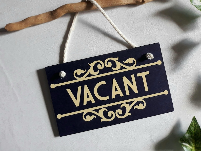 Puede incluir: Un letrero de madera azul marino con la palabra "VACANT" en letras doradas. El letrero tiene adornos dorados decorativos y est&aacute; suspendido por un cord&oacute;n blanco de una rama de madera.
