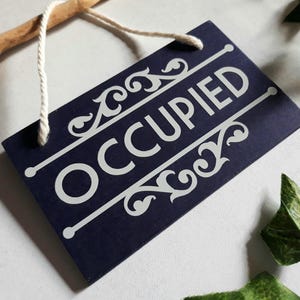 Puede incluir: Un letrero rectangular azul marino con la palabra "OCCUPIED" en blanco, texto estilizado. El letrero tiene adornos blancos decorativos y est&aacute; suspendido por una cuerda blanca. Se ve una rama de madera.