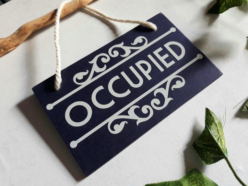 Puede incluir: Un letrero rectangular azul marino con la palabra "OCCUPIED" en blanco, texto estilizado. El letrero tiene adornos blancos decorativos y est&aacute; suspendido por una cuerda blanca. Se ve una rama de madera.