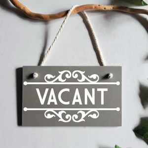 Puede incluir: Un letrero de madera gris con la palabra "VACANT" en blanco, enmarcado por adornos decorativos. El letrero cuelga de una rama natural con una cuerda blanca. Hojas verdes en el fondo.