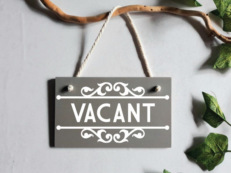 Puede incluir: Un letrero de madera gris con la palabra "VACANT" en blanco, enmarcado por adornos decorativos. El letrero cuelga de una rama natural con una cuerda blanca. Hojas verdes en el fondo.