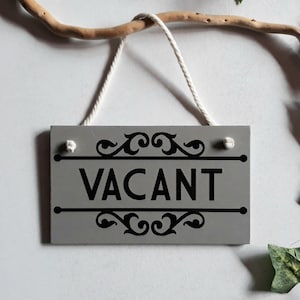 Puede incluir: Un letrero rectangular de madera gris con la palabra "VACANT" en negro, enmarcado por adornos decorativos. El letrero cuelga de una cuerda blanca y est&aacute; suspendido de una rama de madera. Hojas verdes en el fondo.
