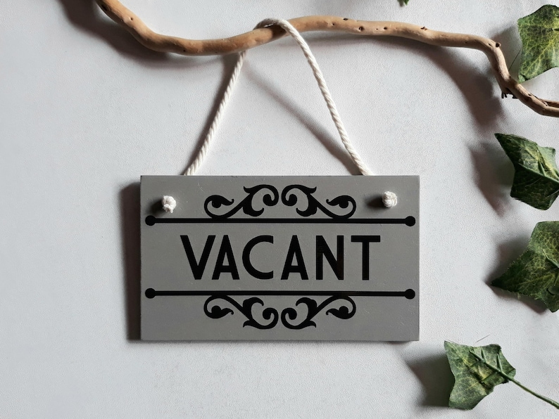 Puede incluir: Un letrero rectangular de madera gris con la palabra "VACANT" en negro, enmarcado por adornos decorativos. El letrero cuelga de una cuerda blanca y est&aacute; suspendido de una rama de madera. Hojas verdes en el fondo.