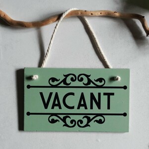 Puede incluir: Un letrero rectangular de madera verde menta con la palabra "VACANT" en letras negras en negrita. El letrero tiene adornos decorativos negros arriba y debajo del texto, y est&aacute; suspendido por una cuerda blanca. Una rama marr&oacute;n est&aacute; encima del letrero.