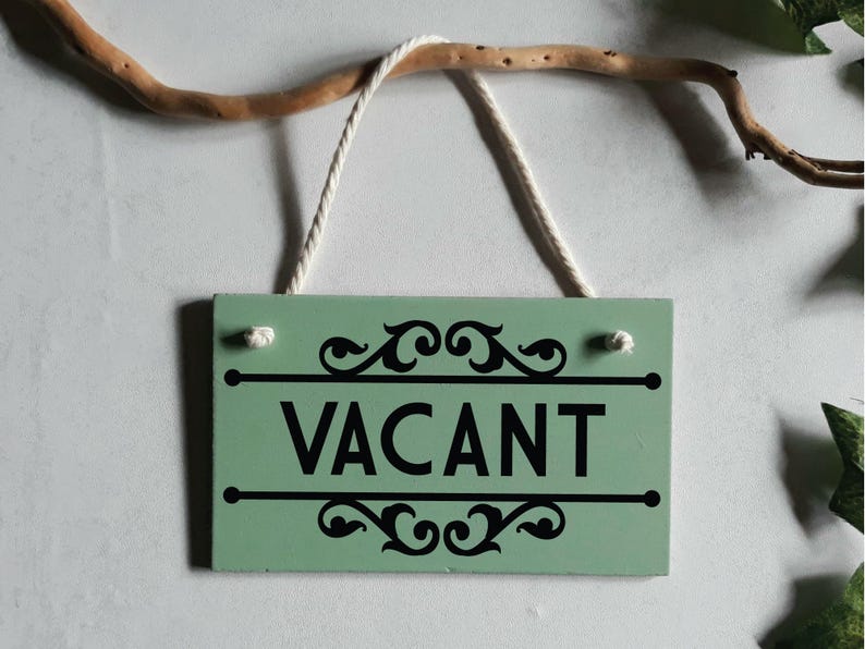 Puede incluir: Un letrero rectangular de madera verde menta con la palabra "VACANT" en letras negras en negrita. El letrero tiene adornos decorativos negros arriba y debajo del texto, y est&aacute; suspendido por una cuerda blanca. Una rama marr&oacute;n est&aacute; encima del letrero.