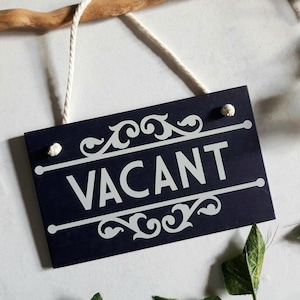 Puede incluir: Un letrero rectangular azul marino con la palabra "VACANT" en blanco, adornado con adornos decorativos. El letrero cuelga de una cuerda blanca y est&aacute; sujeto a una rama de madera. Hojas verdes dispersas alrededor.
