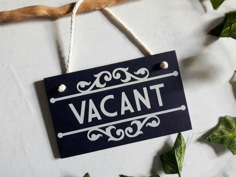 Puede incluir: Un letrero rectangular azul marino con la palabra "VACANT" en blanco, adornado con adornos decorativos. El letrero cuelga de una cuerda blanca y est&aacute; sujeto a una rama de madera. Hojas verdes dispersas alrededor.