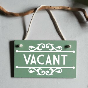 Puede incluir: Un letrero rectangular de madera verde salvia con la palabra "VACANT" en blanco, colgado de una cuerda blanca. El letrero tiene adornos blancos y est&aacute; suspendido de una rama marr&oacute;n.