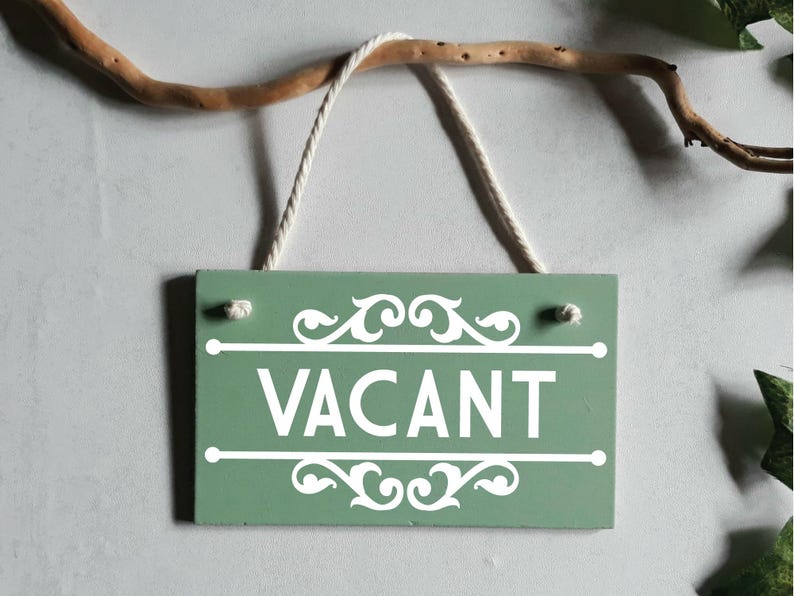 Puede incluir: Un letrero rectangular de madera verde salvia con la palabra "VACANT" en blanco, colgado de una cuerda blanca. El letrero tiene adornos blancos y est&aacute; suspendido de una rama marr&oacute;n.