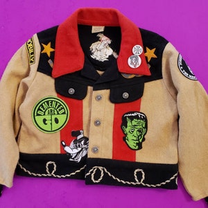 Pode incluir: Uma jaqueta de lã bege, preta e vermelha com uma variedade de patches e pinos. Os patches incluem um círculo verde com as palavras "Demented Are Go", uma ilustração em preto e branco de um monstro e um patch preto e branco com as palavras "The Master". A jaqueta tem um colarinho vermelho e uma borda preta ao redor das bordas.