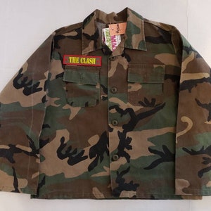 Puede incluir: Una camisa de manga larga con estampado de camuflaje y un parche rojo que dice "THE CLASH" en el pecho izquierdo.