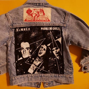 May include: A light blue denim jacket with patches on the back. The patches feature the band names "Bad Brains" and "The Damned" with the album title "Problem Child".