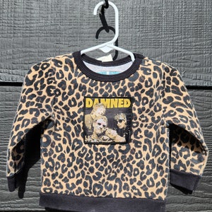 May include: A brown and black leopard print sweatshirt with a black band around the neck and cuffs. The sweatshirt has a black and white patch on the front with the word "DAMNED" in yellow letters and a band with four people on it.