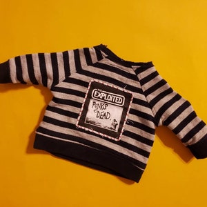 Può includere: Una felpa a righe nere e grigie con una toppa cucita sul davanti. La toppa è nera e bianca e dice "The Exploited" e "Punks Not Dead."