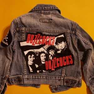 May include: A blue denim jacket with a large patch on the back featuring a band called "Buzzcocks" with a black and white photo of the band members. The patch has red lettering and the band's name is repeated twice. There is a smaller patch on the left sleeve with a skull and crossbones.