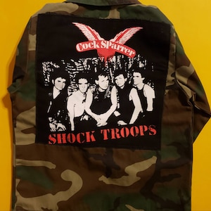 May include: A camouflage jacket with a black patch featuring a red bird with the text "Cock Sparrer" and "Shock Troops" in red letters. The patch also features a black and white image of five people.