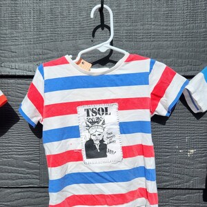 May include: A white, red, and blue striped t-shirt with a patched-on image of a person with the text "TSOL True Sounds of Liberty".