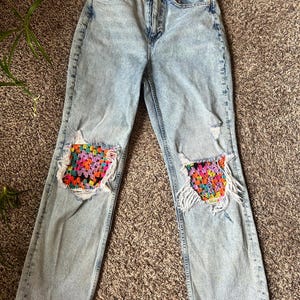 May include: Light wash denim jeans with distressed knees featuring colorful crochet patches. The jeans have a classic five-pocket design and a straight leg fit. The crochet patches showcase a vibrant mix of colors.