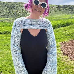 May include: A light blue crochet shrug with long sleeves. The shrug is open in the front and has a crocheted neckline. The model is wearing the shrug over a black tank top.