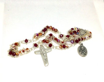 May include: A rosary with red and iridescent beads. The beads are strung on a silver chain. A silver crucifix and a silver pendant with a religious figure are attached to the rosary.