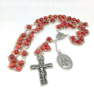 Puede incluir: Un rosario con cuentas rojas iridiscentes y eslabones de cadena plateados. El rosario presenta un crucifijo plateado y un colgante ovalado plateado con una figura religiosa. Las cuentas tienen aproximadamente 6 mm de diámetro.