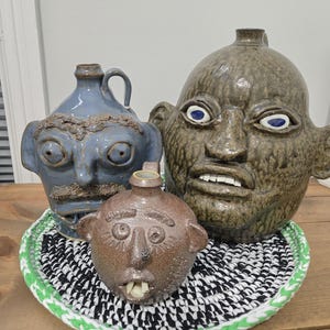May include: Three ceramic face jugs in shades of blue and brown, with exaggerated features. The jugs are arranged on a black and white patterned trivet with a green border, set on a wooden surface.