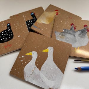 May include: A collection of square kraft paper cards featuring hand-painted illustrations of various birds including white ducks, grey chickens, a yellow hen, and black speckled guinea fowl, all depicted against a backdrop of white painted snow.