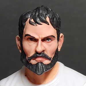Peut inclure: Gros plan sur une tête de figurine d'action en plastique peinte avec une barbe et une moustache noires. La figurine a une expression sérieuse avec les sourcils froncés et un regard déterminé.