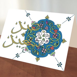 May include: A white card with a blue and gold design. The design features a circular pattern with intricate floral details and Arabic calligraphy that reads "Eid Mubarak".