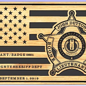 May include: Wooden plaque featuring an American flag design with black stripes and stars. A sheriff's badge is on the right, with text including "John Dutton" and "Boone County Sheriff Dept." The plaque reads "Retired September 1, 2019."