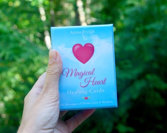 May include: A deck of "Magical Heart Healing Cards" is held against a blurred green background. The card box is light blue with a pink heart and white wings. The text "Magical Heart Healing Cards" is written on the box.