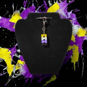 May include: A black key chain with a silver key ring and a yellow, white, and purple charm. The charm is shaped like a rectangular block with two black dots.