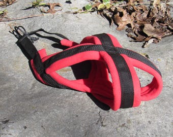 May include: A red and black dog harness. The harness is made of a soft, padded material with black straps that crisscross over the dog's chest and back. It has adjustable straps and a black cord for securing the harness.