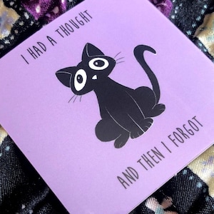 Può includere: Un adesivo quadrato color lavanda con un'illustrazione di un gatto nero e il testo "I HAD A THOUGHT AND THEN I FORGOT". L'adesivo è posto su uno sfondo di tessuto trapuntato scuro con motivi floreali e geometrici.