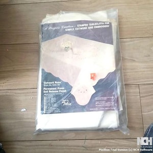 May include: A white tablecloth with a floral design, packaged in plastic. The tablecloth is labeled "A Progress Creation - Stamped Tablecloth for Simple Cutwork and Embroidery." It is also labeled "Cutwork Rose, Style No. 1400/45, Permanent Press, Soil Release Finish."