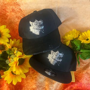 May include: Two black baseball caps featuring a silver embroidered graphic of a snarling wolf head on the front panels. The caps have a structured crown and a flat brim, with one cap showing a distressed brim edge and a circular sticker on the visor.